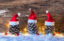 Three pine cones decorated with red santa claus caps in snow with lights and christmas ornaments for christmas by Alex Winter