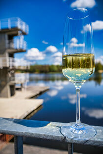 Close-up on a glass of sparkling champagne mirroring the diving platform, blue sky and clouds and the lake in the background by Claudia Schmidt
