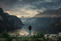 Man standing at waterfront of lake in the mountain landscape Eidfjord fjord in Norway during sunset by Bastian Linder
